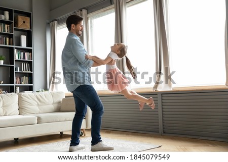 Full length energetic young father spinning laughing joyful little preschool child daughter, having fun together in modern living room. Smiling daddy lifting in air funny small kid girl at home. Royalty-Free Stock Photo #1845079549