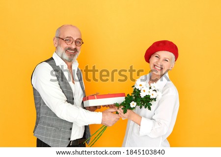 Studio shot of elegant bald unshaven man pensioner in spectacles giving birthday gift to his adorable middle aged wife, posing isolated with box and bunch of daisies, having happy facial expression