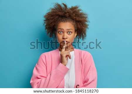 Mysterious dark skinned woman makes silence gesture and looks worried at camera asks not to tell her secret presses index finger to lips as sign to be quiet or mute wears casual pink jumper.