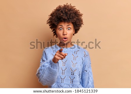 Shocked speechless young Afro American woman stares at camera and points index finger in front wears casual winter blue jumper sees something horrible into distance poses against beige background
