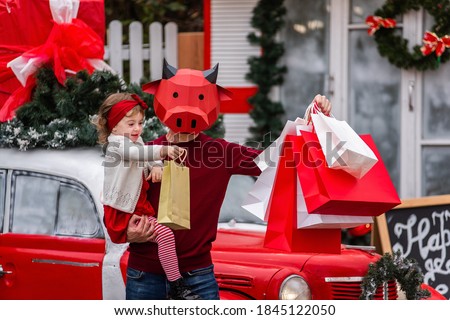 A young man in a red bull mask is holding a little baby, white packages. New Year's shopping, sale. The family stands by the car, trees in the snow decorated for Christmas. Girl kiss an ox. Copy space