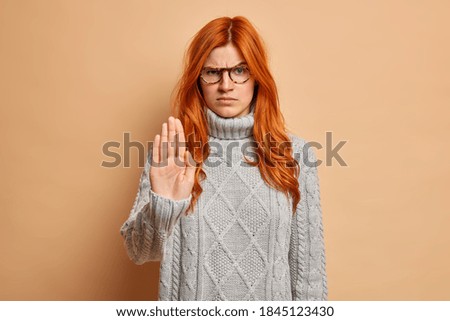 Angry irritated woman with red hair makes stop gesture stretches arm raises eyebrows and looks with grumpy expression wears winter sweater shows prohibition sign isolated over beige background