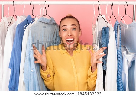 Shopaholic Concept. Portrait Of Surprised Emotional Lady Standing At Clothing Rail In Closet. Screaming Young Woman Shouting And Gesturing With Joy, Choosing Clothes Over Pastel Pink Studio Background