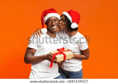 Happy black woman giving her boyfriend Christmas gift on orange studio background, copy space. Cheerful african american family in Santa hats celebrating New Year together, lady kissing man