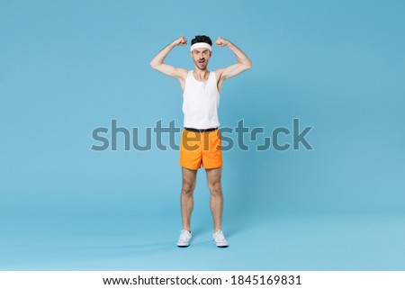Full length portrait of shocked strong young fitness man with skinny body sportsman in headband shirt shorts showing biceps muscles isolated on blue background. Workout gym sport motivation concept Royalty-Free Stock Photo #1845169831