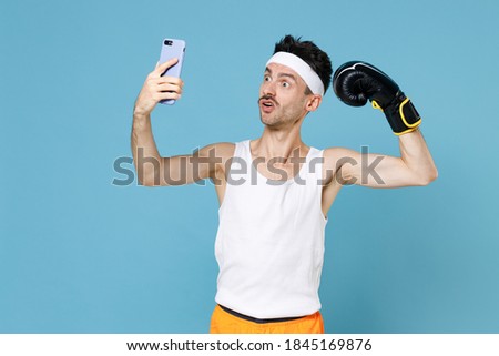 Shocked man boxer with skinny body sportsman in headband shirt shorts boxing gloves doing selfie shot on mobile phone showing biceps muscles isolated on blue background. Workout gym sport concept