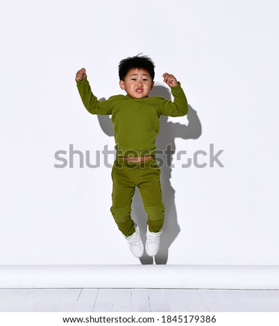 Faceless dark-haired boy jumping against the background of a white wall in the studio. The child is dressed in a stylish green tracksuit and white sneakers.