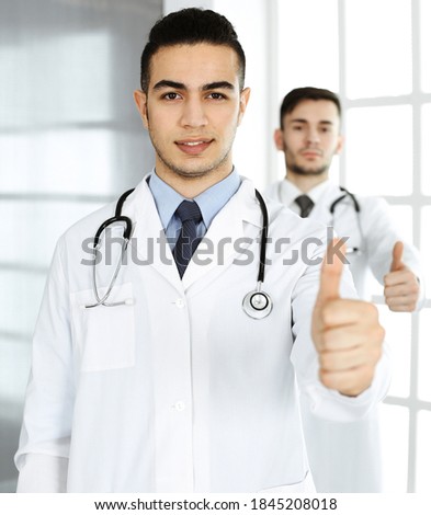 Arab doctor man showing Ok sign with thumbs up with caucasian colleague in medical office or clinic. Diverse doctors team, best treatment, medicine and healthcare concept