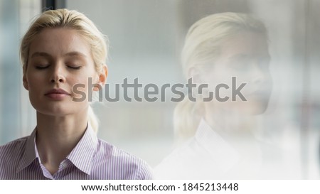 Head shot close up mindful young businesswoman breathing fresh air with closed eyes near window, enjoying calm break pause time in office, reducing stress pressure during workday, copy space for text. Royalty-Free Stock Photo #1845213448