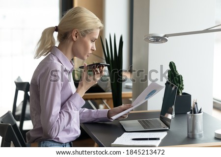 Concentrated smart young blonde businesswoman reading paper marketing research report, holding loudspeaker cell phone call conversation with colleagues, discussing results or development strategy.