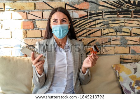 Pretty young woman with protective face mask using mobile phone anc credit card in the cafe