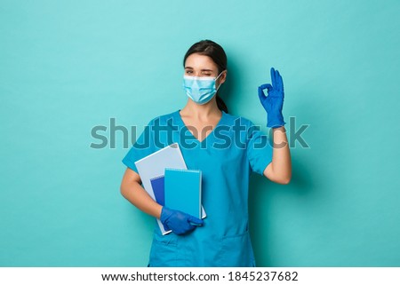 Covid-19, pandemic and medicine concept. Image of confident, beautiful female doctor in medical mask, gloves and scrubs, holding notebooks and showing okay sign, blue background
