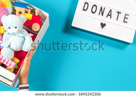 Hands holding donation box with toys books clothing and lightbox with text Donate on light blue background. Top view