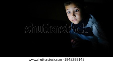 Young boy covered her mouth in fright, sitting at a computer on a dark night. The child is shocked by what he saw on the laptop. Watching a scary movie.