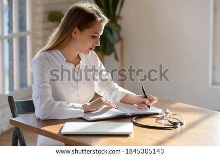 Doctor at paperwork. Responsible concentrated millennial woman family therapist sitting at desk in hospital cabinet making recordings to paper casebook, writing medical history or treatment profile Royalty-Free Stock Photo #1845305143