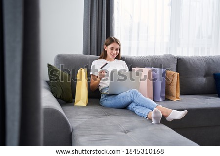 Cheerful smiling lady sitting at the sofa in her modern flat and using a laptop while having online shopping with her credit card. Stock photo