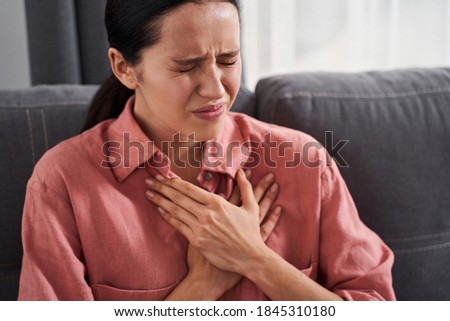 Caucasian woman wearing pink shirt having a severe chest pain while sitting on the sofa in her home. Heart attack, infarction concept Royalty-Free Stock Photo #1845310180