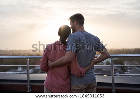 Waist up portrait of beautiful young couple embracing and looking at the sun while standing at the balcony. Romantic couple spending time together with love during sunset Royalty-Free Stock Photo #1845311053