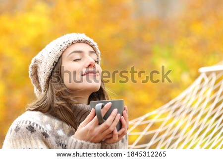 Relaxed woman breaths fresh air holding coffee cup for breakfast sitting on a rope hammock in autumn in a forest Royalty-Free Stock Photo #1845312265