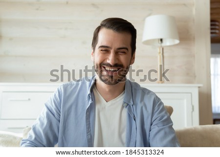 Glad to see you! Profile portrait picture of handsome young man sitting on couch at home alone making videocall by electronic gadget looking at camera greeting dialogue partner laughing on good joke