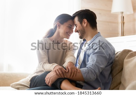 Happy together. Smiling newlyweds cuddling on couch at living room of rented purchased flat, caring husband hugging holding on knees beloved wife, young couple is glad to start new independent life Royalty-Free Stock Photo #1845313660