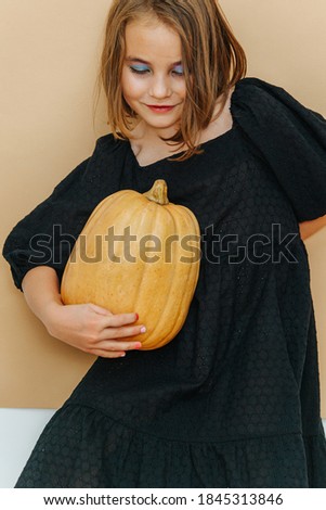 Halloween girl in a black dress, holding pumpkin. Simplisitc halloween. She has a heavy make up, blue eye shadows and a lipstick. Over brown. Assuming stylish pose, looking down.