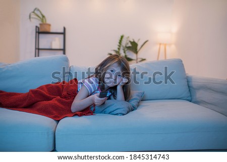 Bored little girl watching televesion with a sceptical bored face expression, leaning on her hand under a blanket. Holding remote.