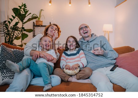 Laughing family of parents and two children watching comedy together on the couch in the living room.