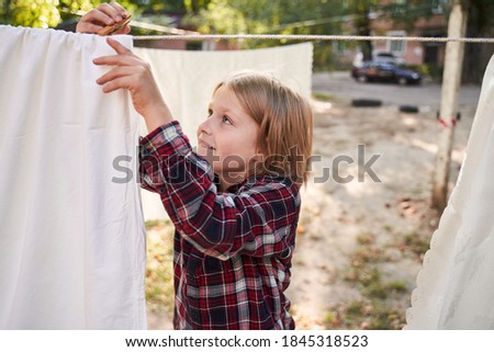 Grandma's helper. Photo of concentrated blonde little boy diligently attaching clothes with clothespins to dry them outdoors near the country house