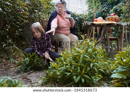 Senior careful grandmother giving advice to her grandson how best to weeding the garden while he helping her in the beds. Stock photo. Family support and helping concept