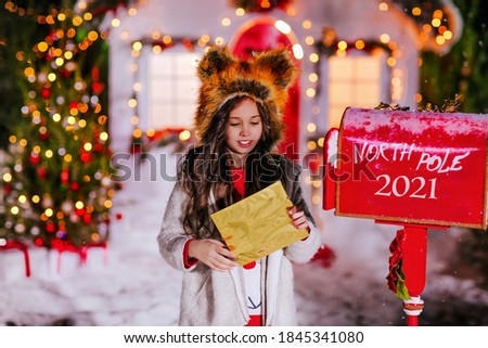 A beautiful girl in red dog hat sends a letter to Santa in a red mailbox on the background of a Christmas decorated house. High quality photo