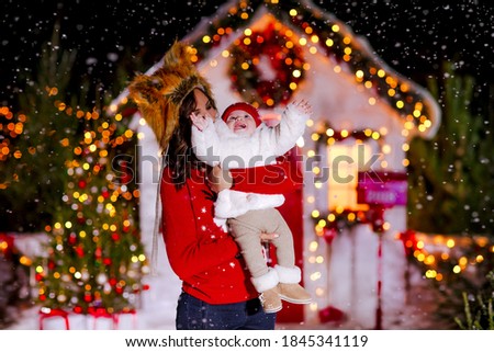 A mother in a fur red fox hat holds a small child in her arms against the backdrop of a Christmas house. Copy space