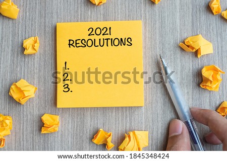 2021 RESOLUTIONS word on yellow note with Businessman holding pen and crumbled paper on wooden table background. New Year start, Strategy and Goal concept