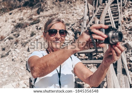Elderly senior traveling backpacker mature woman tourist walking taking photos on coast background of sea, stones, rocks, blue sky. Retired people summer holiday vacation, active lifestyle concept