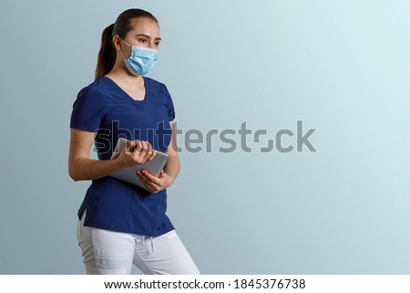 Latina woman, wearing a nurse's uniform with mask, holding a digital tablet in her hands, and a blue background with space for text