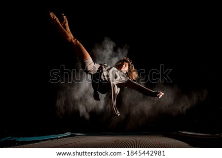young attractive woman jumping while lying on large trampoline at dark time and light of searchlight illuminates her. Some smoke around
