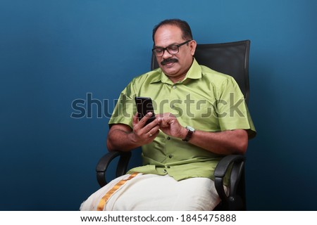 Middle aged man of Indian origin sitting on a chair  checking his mobile phone Royalty-Free Stock Photo #1845475888