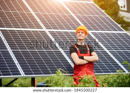 Proud young worker smiling satisfied with his successful work with his arms crossed, on background solar station. Concept: renewable energy, technology, electricity, service, green power.