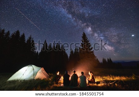 Evening summer camping, spruce forest on background, sky with falling stars and milky way. Group of five friends sitting together around campfire in mountains, enjoying fresh air near illuminated tent Royalty-Free Stock Photo #1845501754