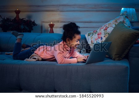 Happy african-american little girl during video call with laptop and home devices, looks delighted and happy. Talking to Santa before New Year's eve, her family, watching cartoons, typing text.