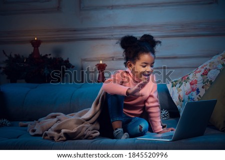 Happy african-american little girl during video call with laptop and home devices, looks delighted and happy. Talking to Santa before New Year's eve, her family, watching cartoons. Holidays miracle.