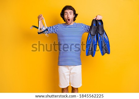 Photo of surprised guy hold scuba diving fins breathing mask goggles wear blue white shorts isolated on yellow color background