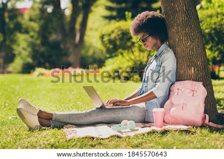 Full length body size side profile photo of black skinned woman with curly hair working on laptop browsing internet in city park