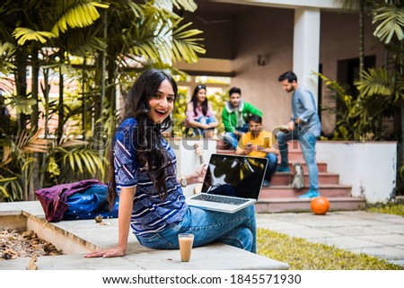 Asian Indian college student in focus working on laptop or reading book while other classmates in the background, outdoor picture in university campus