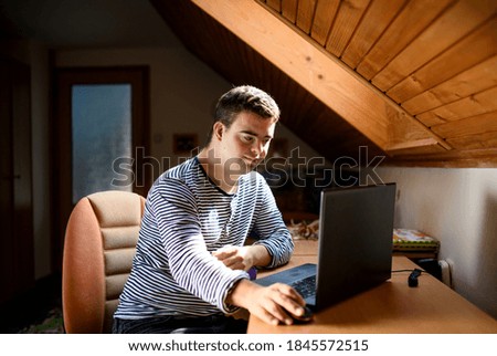 Down syndrome adult man sitting indoors in bedroom at home, using laptop. Royalty-Free Stock Photo #1845572515