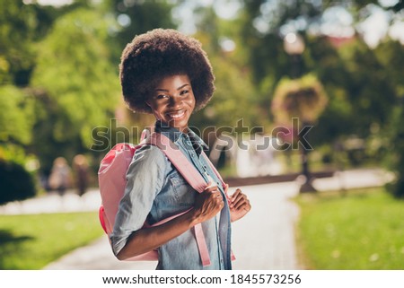 Side profile photo of black skinned pretty girl with curly hair smiling cheerfully keeping bag on back wearing stylish casual denim shirt