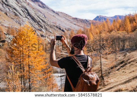 Young guy with glasses and a beard is taking a horizontal picture with his phone while walking on the mountain