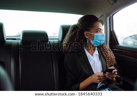 Relaxed woman passenger  in protective medical mask in the taxi car on a backseat with phone. Work on the way to the office. Covid-2019. Royalty-Free Stock Photo #1845640333