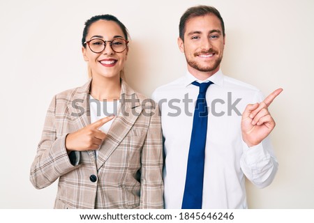 Beautiful couple wearing business clothes with a big smile on face, pointing with hand finger to the side looking at the camera. 
