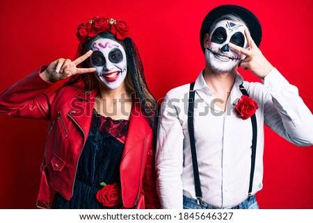 Couple wearing day of the dead costume over red doing peace symbol with fingers over face, smiling cheerful showing victory 